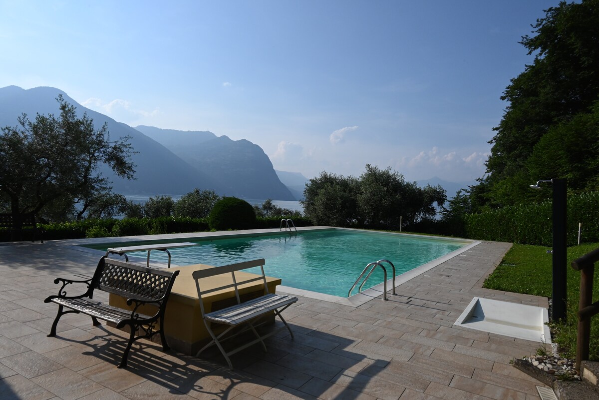 A serene outdoor pool area is surrounded by landscaped greenery and trees, with mountain views in the background. Two lounge chairs are situated beside the pool, which features a gentle gradient of color in the water. The pathway is paved with light-colored tiles.