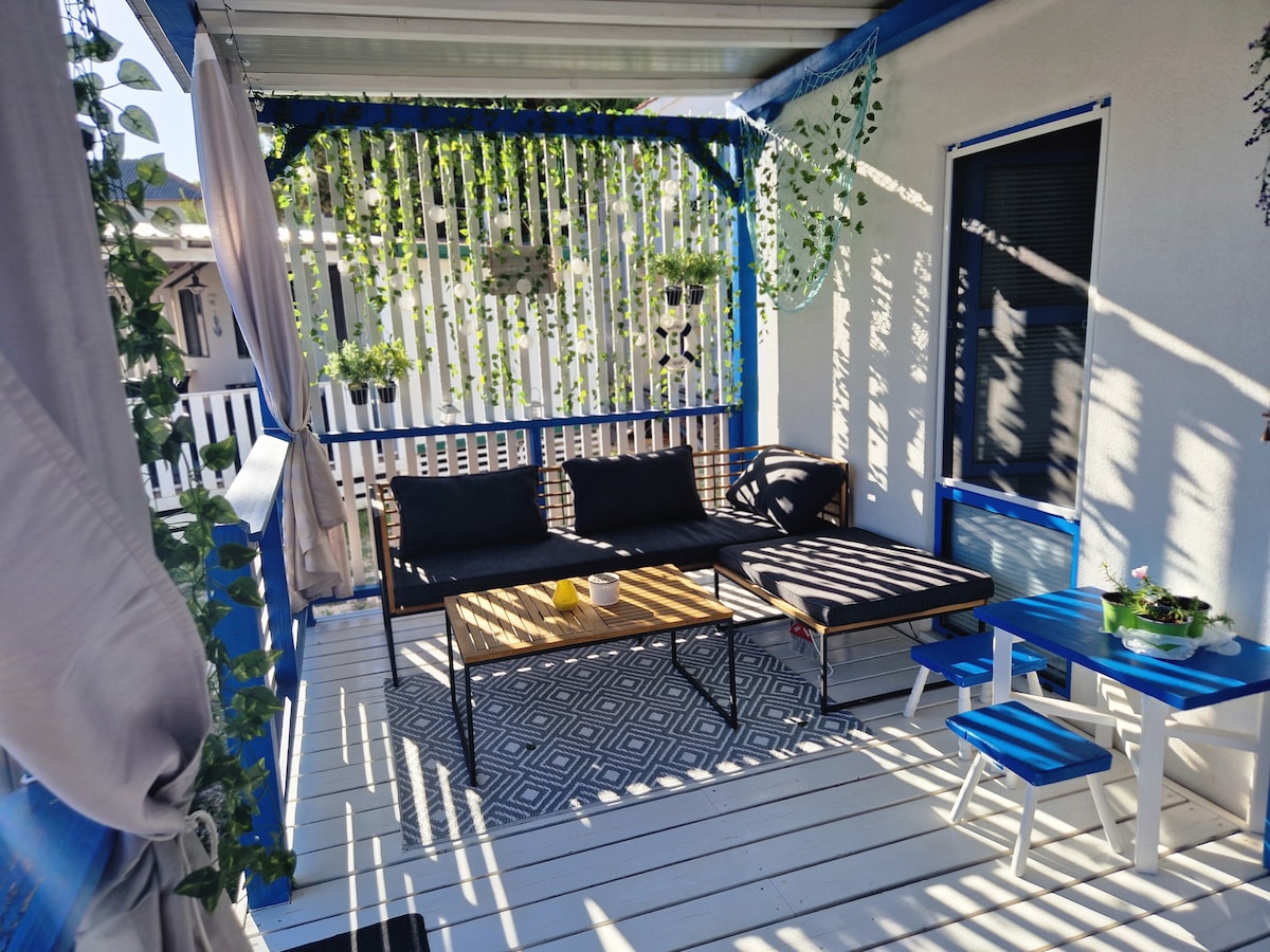 A covered terrace features a cozy seating area with a dark sofa and a coffee table. A small blue table adds color, surrounded by decorative plants. Sunlight filters through the slatted walls, creating a patterned play of light and shadow across the white floor.