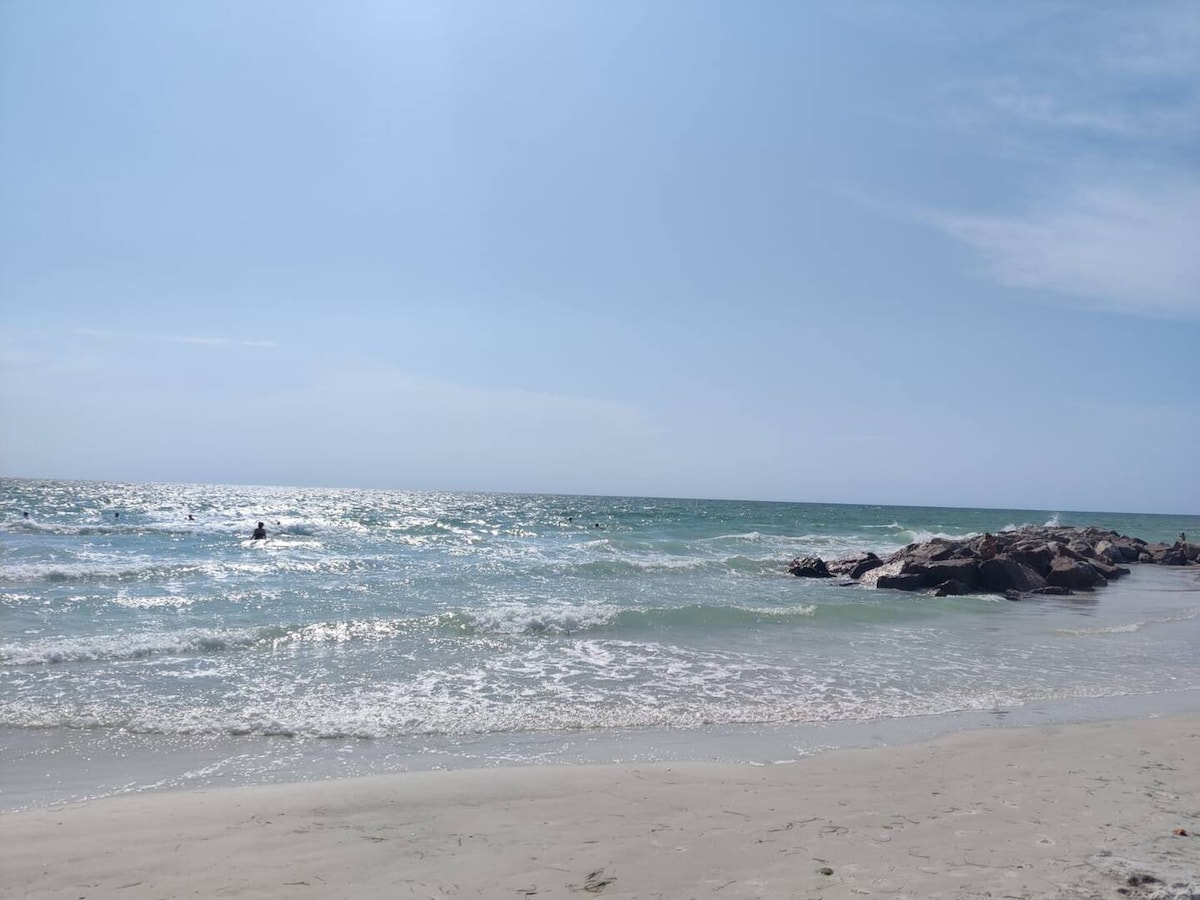 A serene beach scene captures gentle waves lapping against the shore under a clear blue sky. Sunlight reflects off the water, creating a sparkling effect. In the distance, a figure can be seen enjoying the water while rocky formations punctuate the coastline.