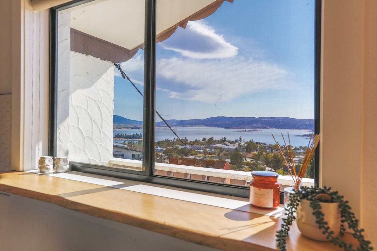 A large window frames a stunning view of Jindabyne Lake and surrounding mountains. A wooden windowsill displays glass candles and a small potted plant, complementing the serene landscape visible beyond. Soft clouds drift across the blue sky, enhancing the tranquil atmosphere.