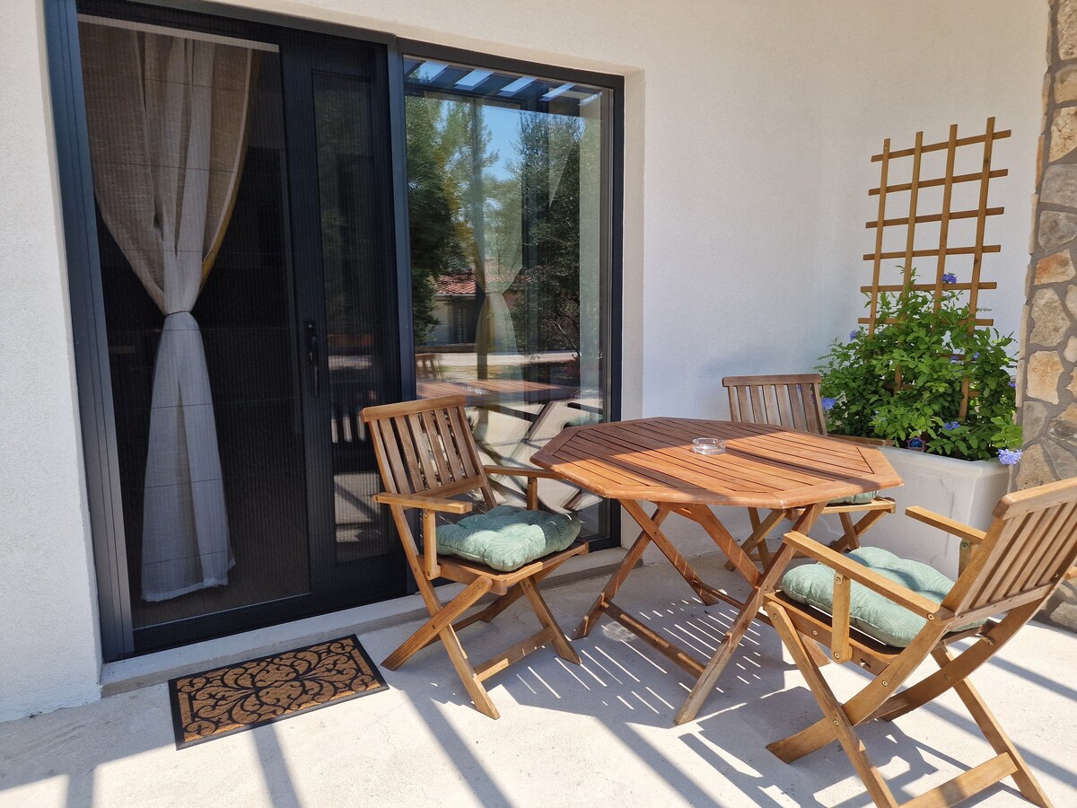 A terrace space is presented with a wooden table surrounded by four folding chairs, each with soft cushions. Natural light filters through the door and a curtain drapes beside it. A decorative trellis with greenery complements the tranquil setting.