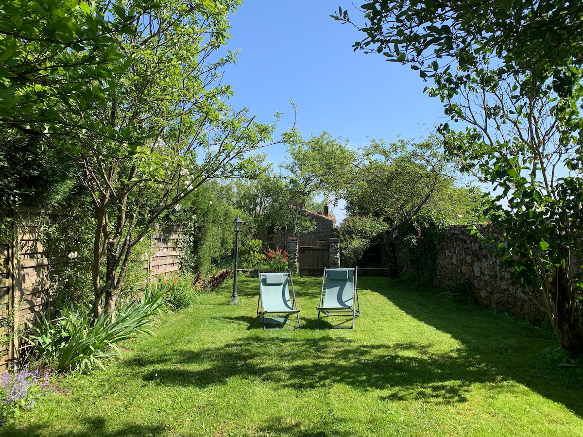 A serene garden space is presented, featuring well-maintained grass and two folding chairs positioned for relaxation. Surrounding trees and flowering plants provide natural shade, while a stone wall creates a sense of privacy. The clear blue sky enhances the peaceful outdoor atmosphere.