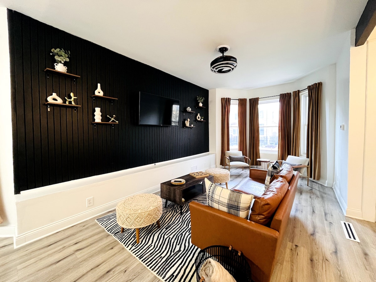 The living area showcases a large leather sofa complemented by a patterned rug. A custom black wooden slat wall serves as a backdrop for a flat-screen TV mounted above. Natural light enters through the expansive window, framed by elegant curtains. Decorative shelves display various curated items.