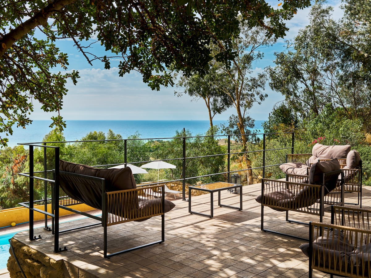 A spacious terrace offers comfortable seating with two chairs and a coffee table, framed by lush greenery and a panoramic view of the azure sea. Sun loungers and umbrellas are positioned nearby, inviting relaxation in a serene outdoor setting.