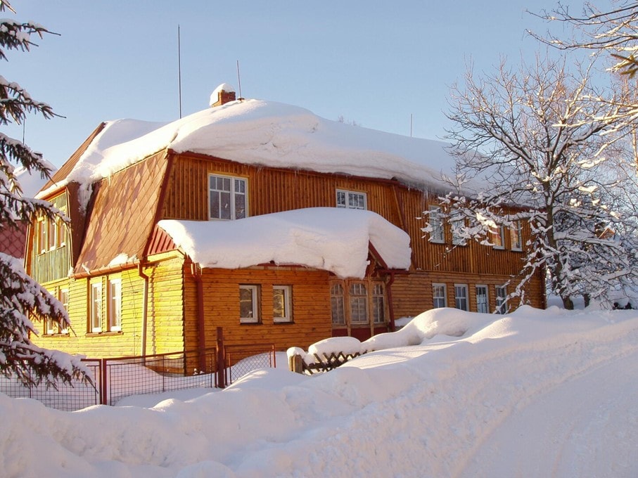 A charming wooden chalet is covered in a blanket of fresh snow, showcasing its sloped roof and large windows. Surrounding trees are also dusted with snow, creating a serene winter scene. A snow-covered path leads up to the inviting entrance.
