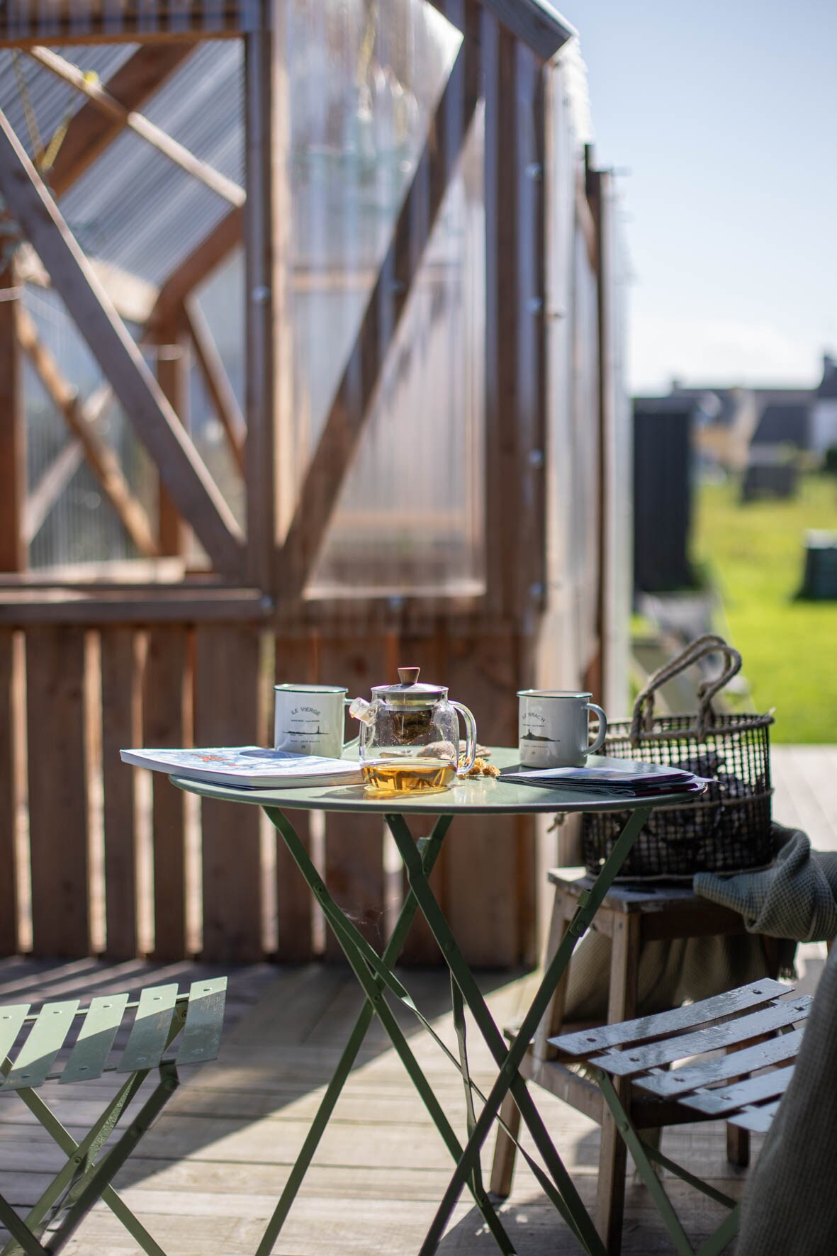 A small outdoor table is set with a glass teapot and two mugs. A magazine lies open on the table, surrounded by light green folding chairs and a wooden bench. Natural light highlights the nearby wooden structure and deck area, offering a welcoming atmosphere.