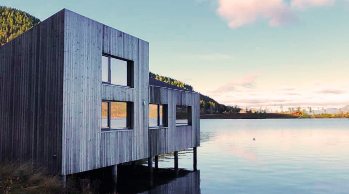 A contemporary wooden cabin is shown standing on stilts above a calm body of water. Large windows offer scenic views of the surrounding landscape, while a tranquil sky reflects softly on the water's surface, enhanced by the natural hues of the nearby mountains.