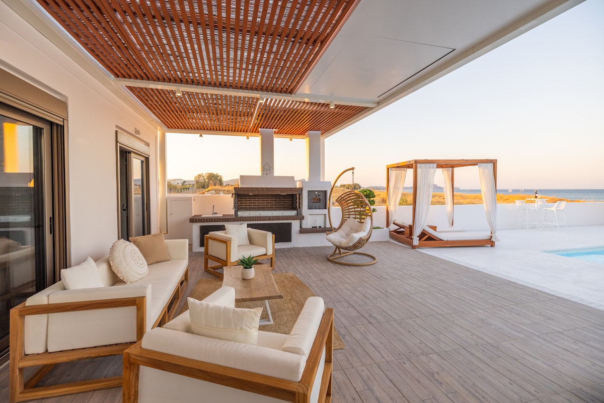 An outdoor living space is presented, featuring a comfortable seating arrangement with wooden frames and soft cushions. A nearby outdoor kitchen is equipped for cooking, while a shaded area includes a cozy hammock. The view stretches towards the ocean, enhancing the serene atmosphere.