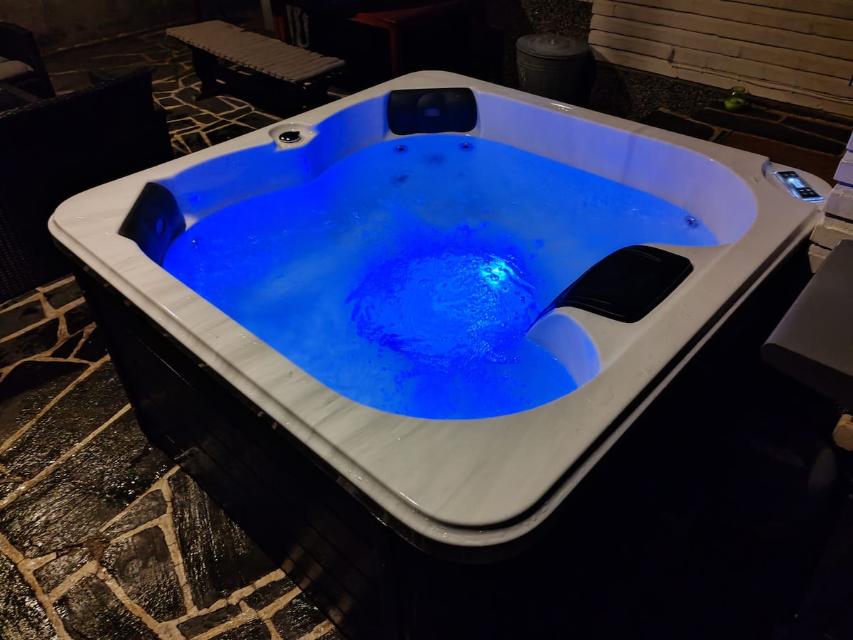 A spacious outdoor hot tub is illuminated in calming blue lights, creating a relaxing ambiance. The tub features comfortable seating and ergonomic design, surrounded by stone flooring, adding to the tranquil outdoor setting.