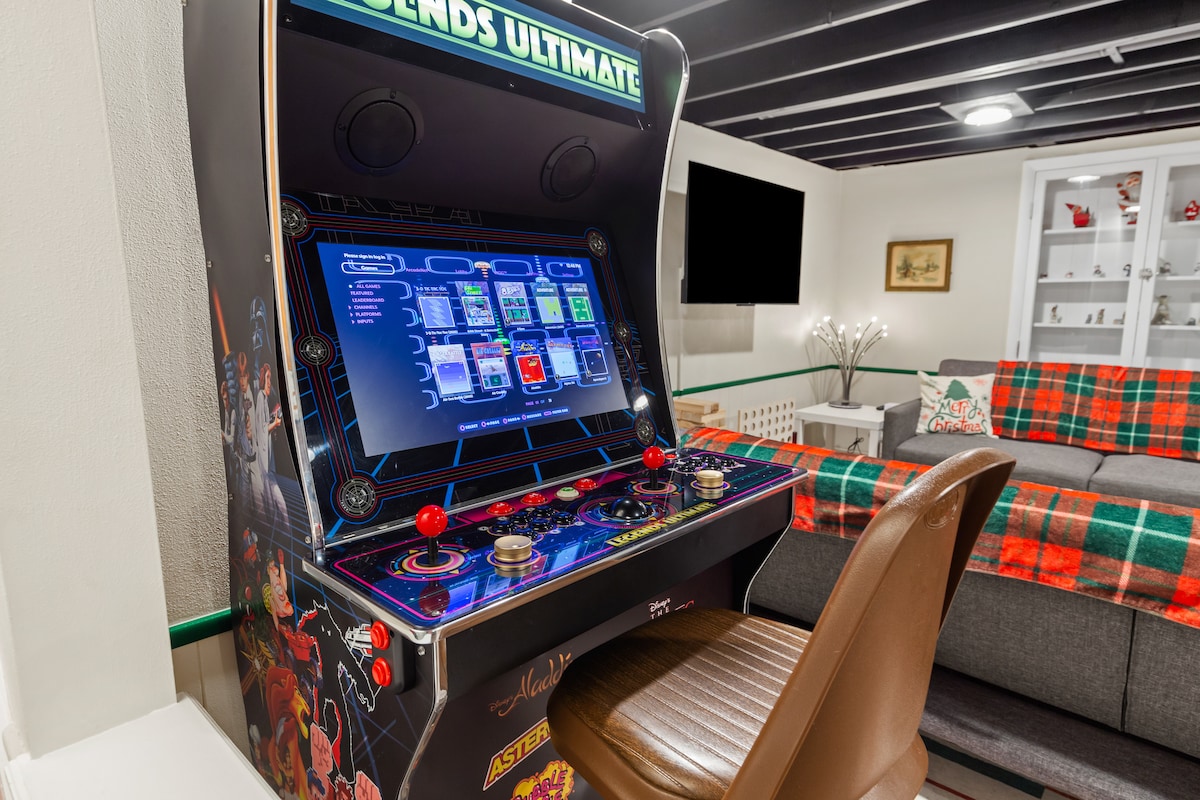 An arcade gaming machine stands against a backdrop of a cozy living area. The machine displays colorful graphics and buttons, inviting interaction. A brown leather chair is positioned in front, and a gray sectional couch with a festive plaid throw is visible in the background.