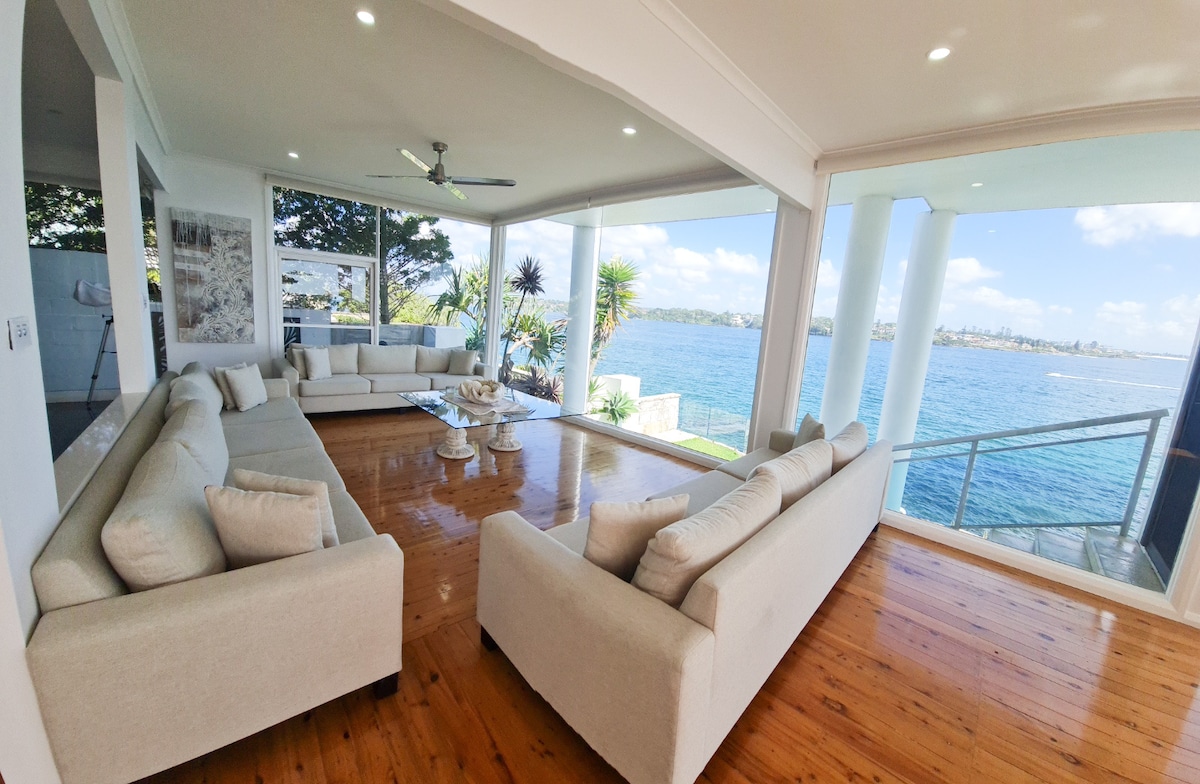 A spacious living area features large glass windows offering views of the ocean. Comfortable couches are arranged around a central coffee table, and the wooden floor adds warmth to the setting. Natural light fills the room, creating a bright and airy atmosphere with direct access to the outdoor space.