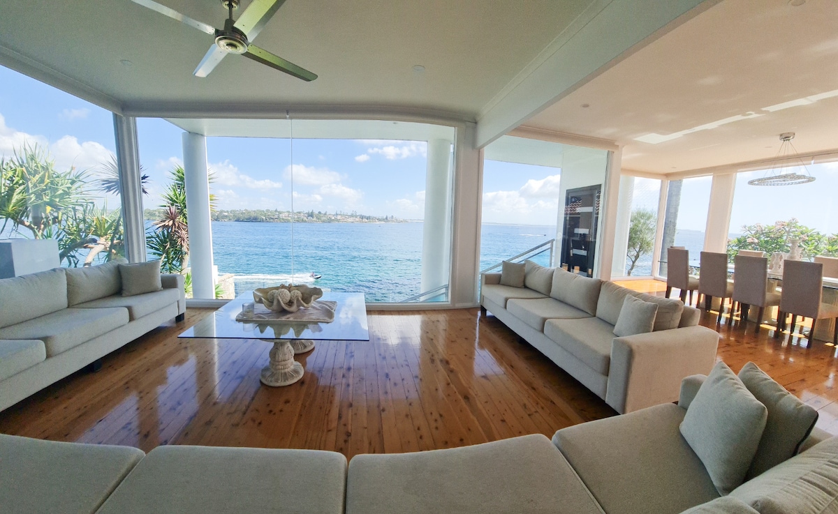 A spacious living area is filled with natural light, offering panoramic ocean views through large glass windows. Two white sofas surround a coffee table at the center. A dining area is visible in the background, featuring a long table surrounded by several chairs.