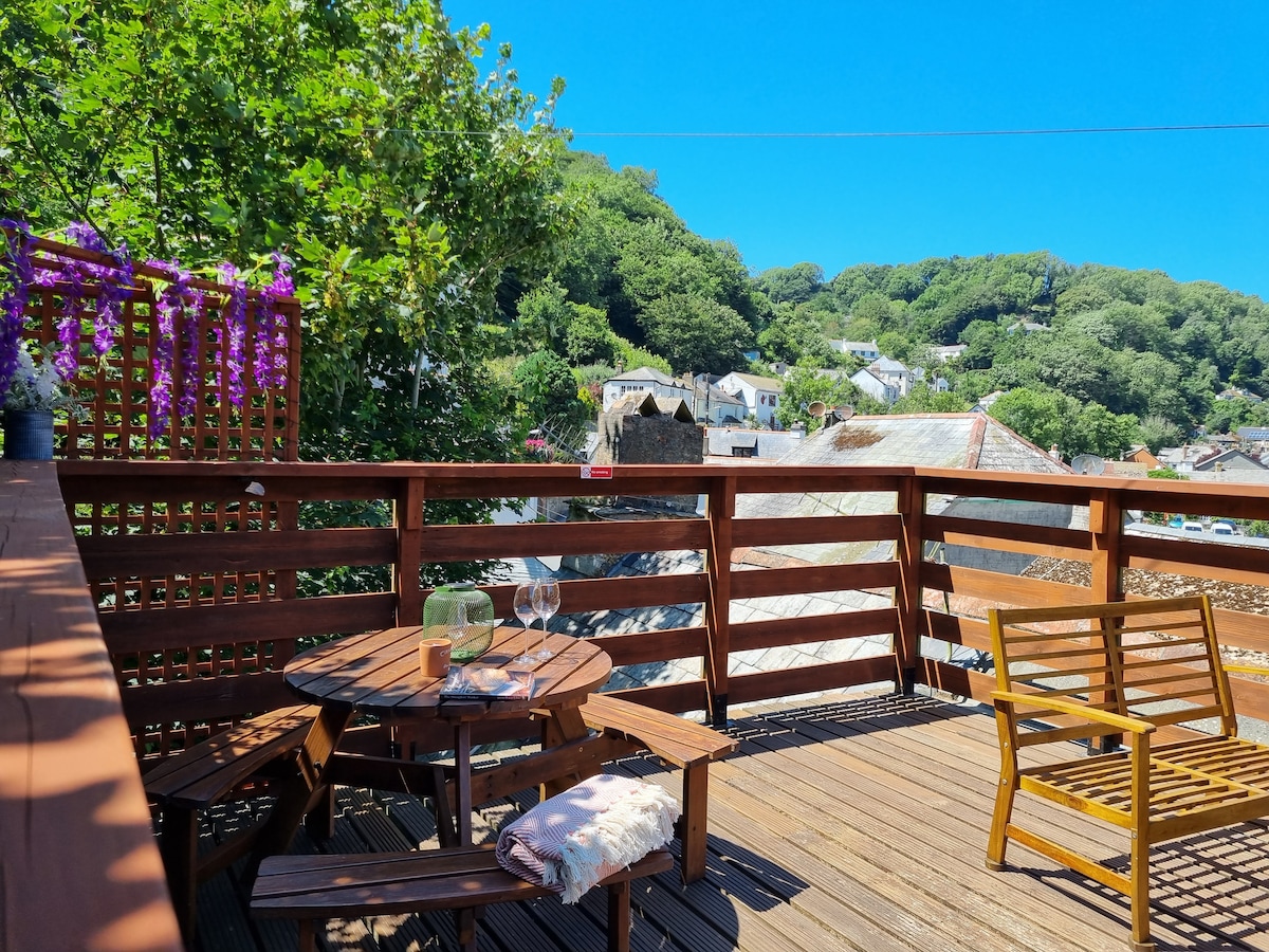 A sunny roof terrace features wooden decking and a table with two glasses, ideal for outdoor relaxation. A wooden chair is placed beside the table, surrounded by greenery. Views of the village rooftops and the hillside create a serene backdrop.