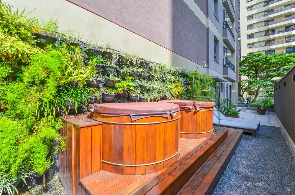 A wooden hot tub is situated on a spacious wooden deck, surrounded by lush greenery and vertical planters. Smooth pebbles create a pathway, enhancing the tranquil outdoor space, while an area for relaxation with lounge chairs is visible in the background.