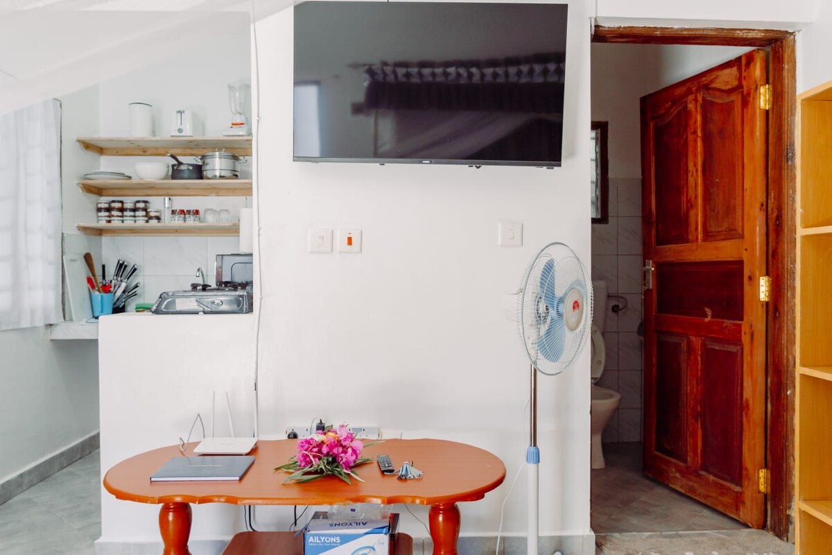 A functional living area is shown, featuring a wooden table adorned with a decorative flower arrangement. A wall-mounted television is visible, and a ceiling fan is positioned nearby. The kitchenette is neatly organized in the background, displaying essential cooking utensils and appliances.