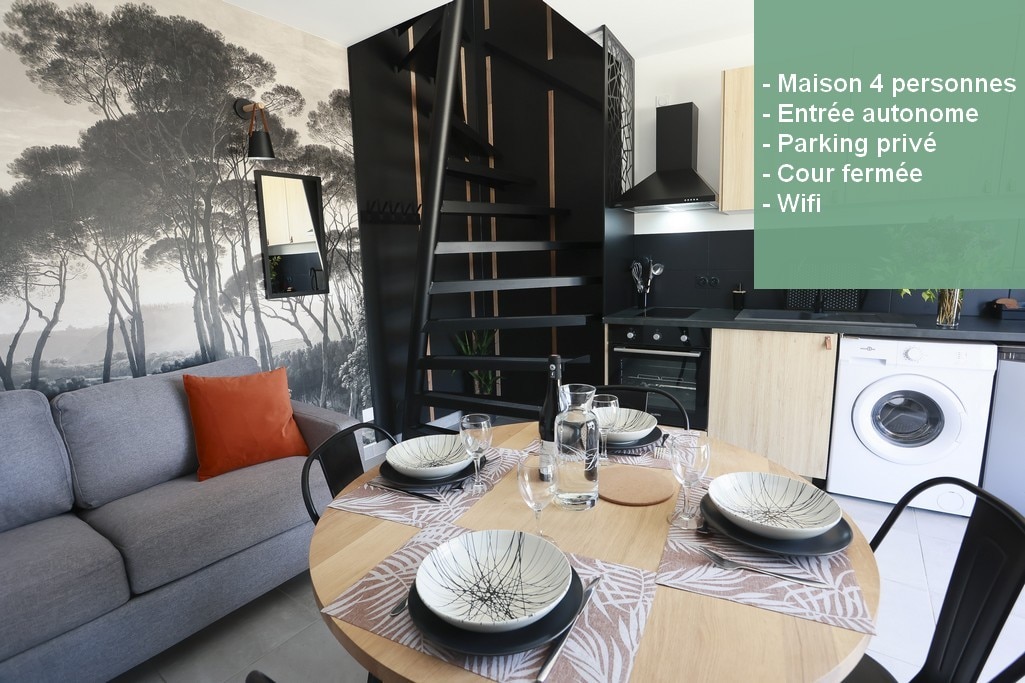 The dining area features a modern table set for four, paired with simple black chairs. A cozy couch is in the background, and a spiral staircase leads to the upper level. The kitchen, adjacent to the dining area, includes contemporary appliances.