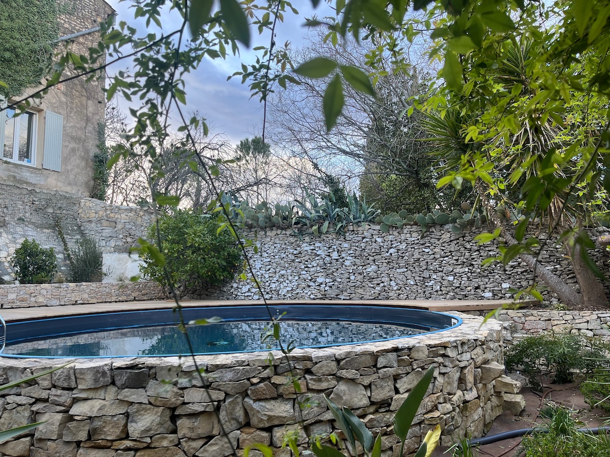 A circular swimming pool is surrounded by a natural stone wall, nestled within a lush garden. Various plants and trees offer shade and privacy, creating a serene outdoor environment. The tranquil water reflects the sky, enhanced by the greenery surrounding the area.