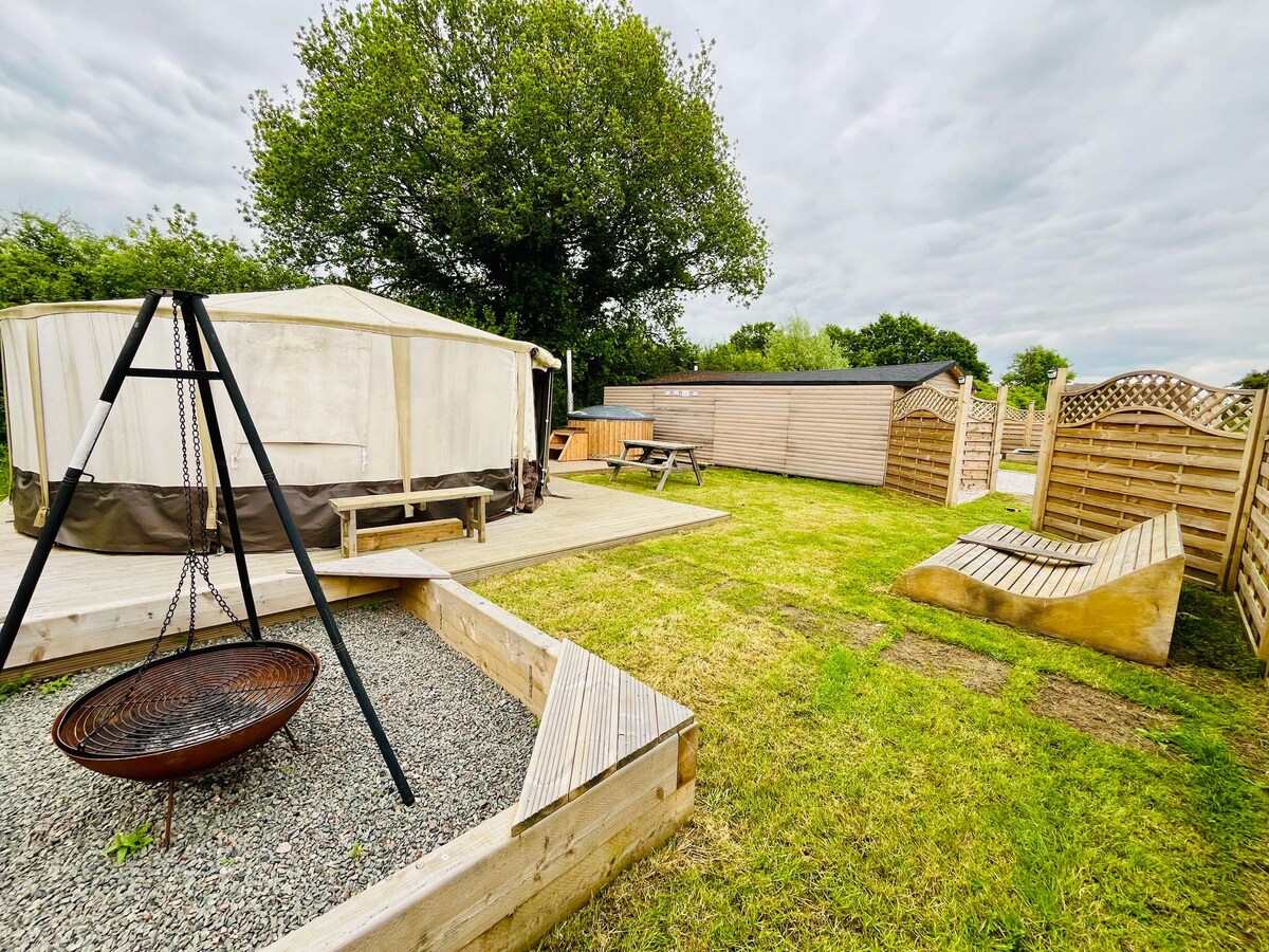The image showcases a grassy area featuring a circular firepit on a gravel base, surrounded by wooden benches. A yurt with a light-colored exterior is visible, along with wooden picnic tables and a fenced garden space with a tree in the background.