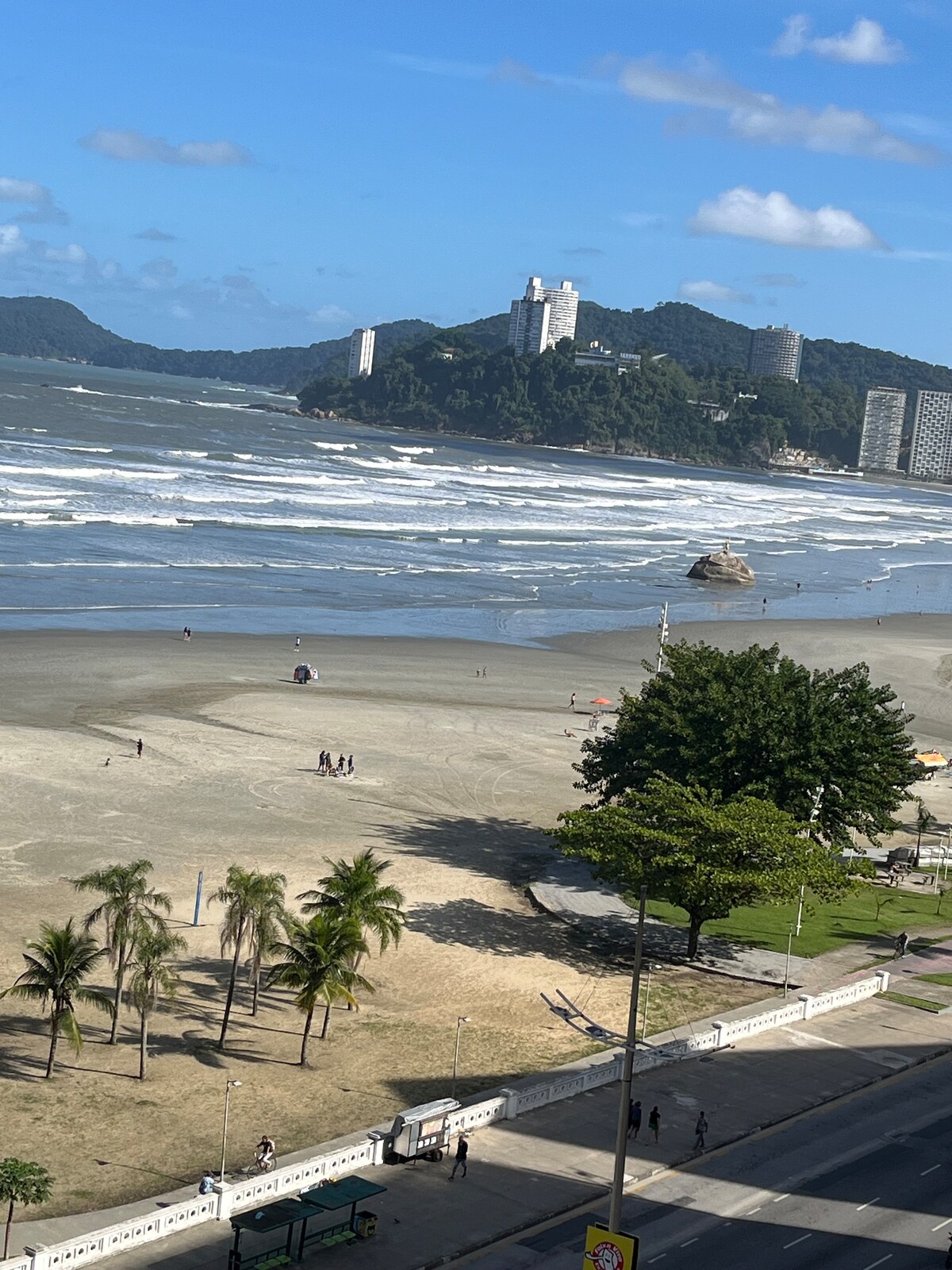 A wide view of a beach showcases gentle waves rolling onto the shore. People can be seen enjoying the sand, with green trees lining the beachfront. In the background, a hill is adorned with tall buildings, contributing to the scenic coastal landscape.