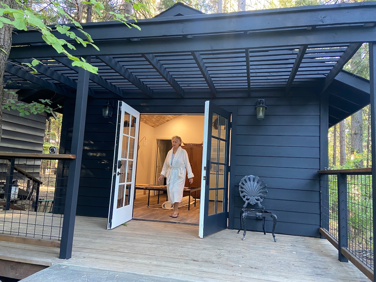 A cozy entrance area is framed by a black exterior with large glass doors, inviting guests inside. A wooden deck features a chair, surrounded by lush trees. Inside, warm light spills from a spacious room, hinting at a welcoming atmosphere.
