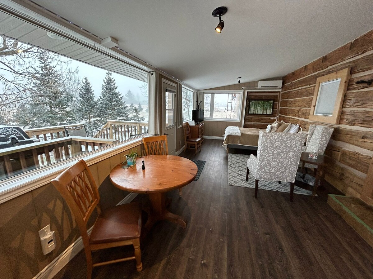 A bright room features large windows allowing natural light to illuminate the space. A round wooden table with two chairs is positioned near the window, while a cozy seating area is arranged with patterned armchairs and a small table, creating an inviting atmosphere.