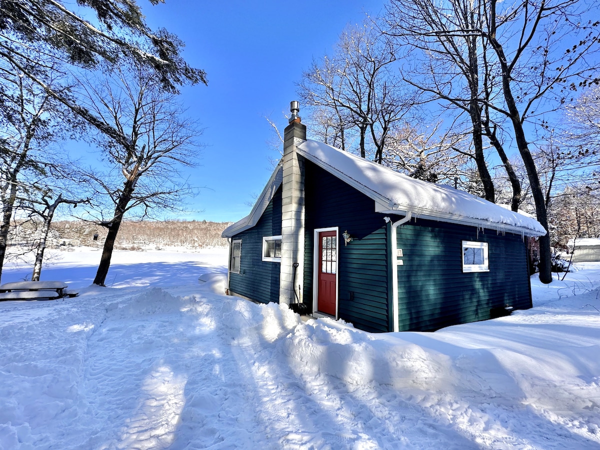 A quaint blue cabin is framed by snow-covered trees, standing beside a frozen lake under a clear blue sky. A red door adds a pop of color to the exterior, while the sunlight casts soft shadows on the snowy ground.