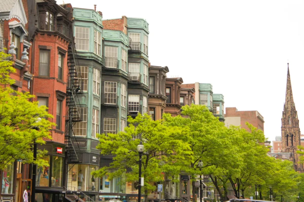 A row of historic brownstone buildings is lined along a tree-filled street. Large windows display architectural details, while lush green trees provide a natural aesthetic. A steeple of a church can be seen in the distance, adding to the city's skyline.