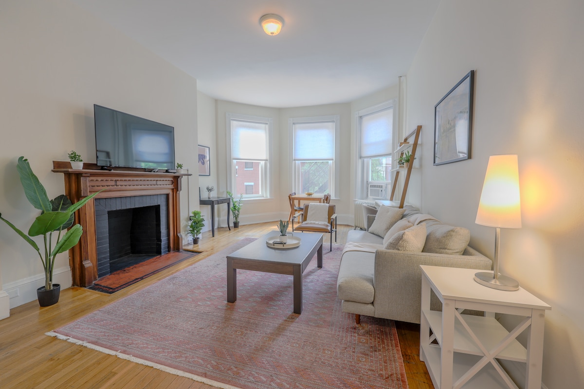 A light-filled living area features a comfortable sofa and a coffee table centered on a large area rug. A fireplace with a decorative mantel is present, accompanied by a flat-screen TV and greenery throughout. Large windows allow ample natural light into the space.