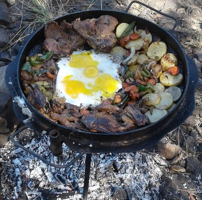 A large, round cooking pan is positioned over a bed of embers, featuring a variety of grilled meats, colorful vegetables, and eggs. The meats and vegetables are arranged around a central circle of eggs, showcasing a rustic outdoor cooking scene.