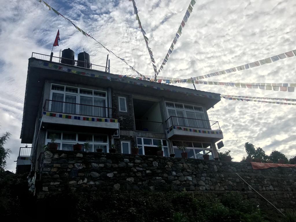 The two-story building is constructed from stone and features large windows on each floor, allowing ample natural light. Colorful prayer flags are displayed above, adding vibrancy to the exterior. A stone wall supports the structure, enhancing its connection with the surrounding landscape.