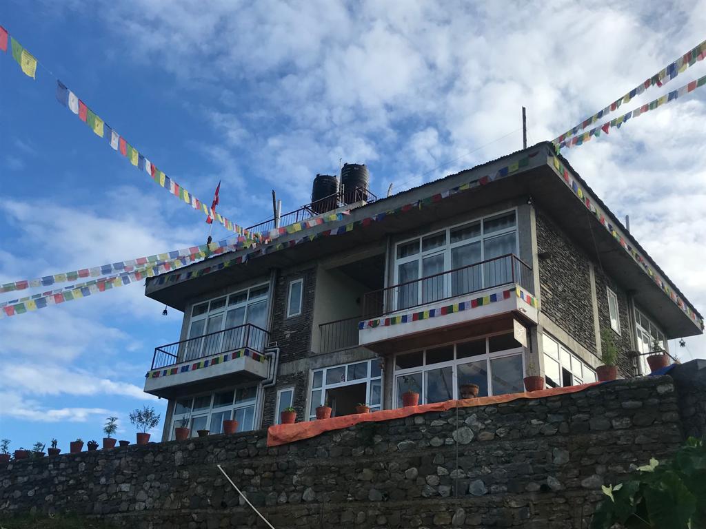 The two-story resort building features large glass windows and balconies, accented by colorful prayer flags. A stone wall forms the base of the structure, with flower pots visible along the edge. The sky is partly cloudy, showcasing a serene atmosphere.