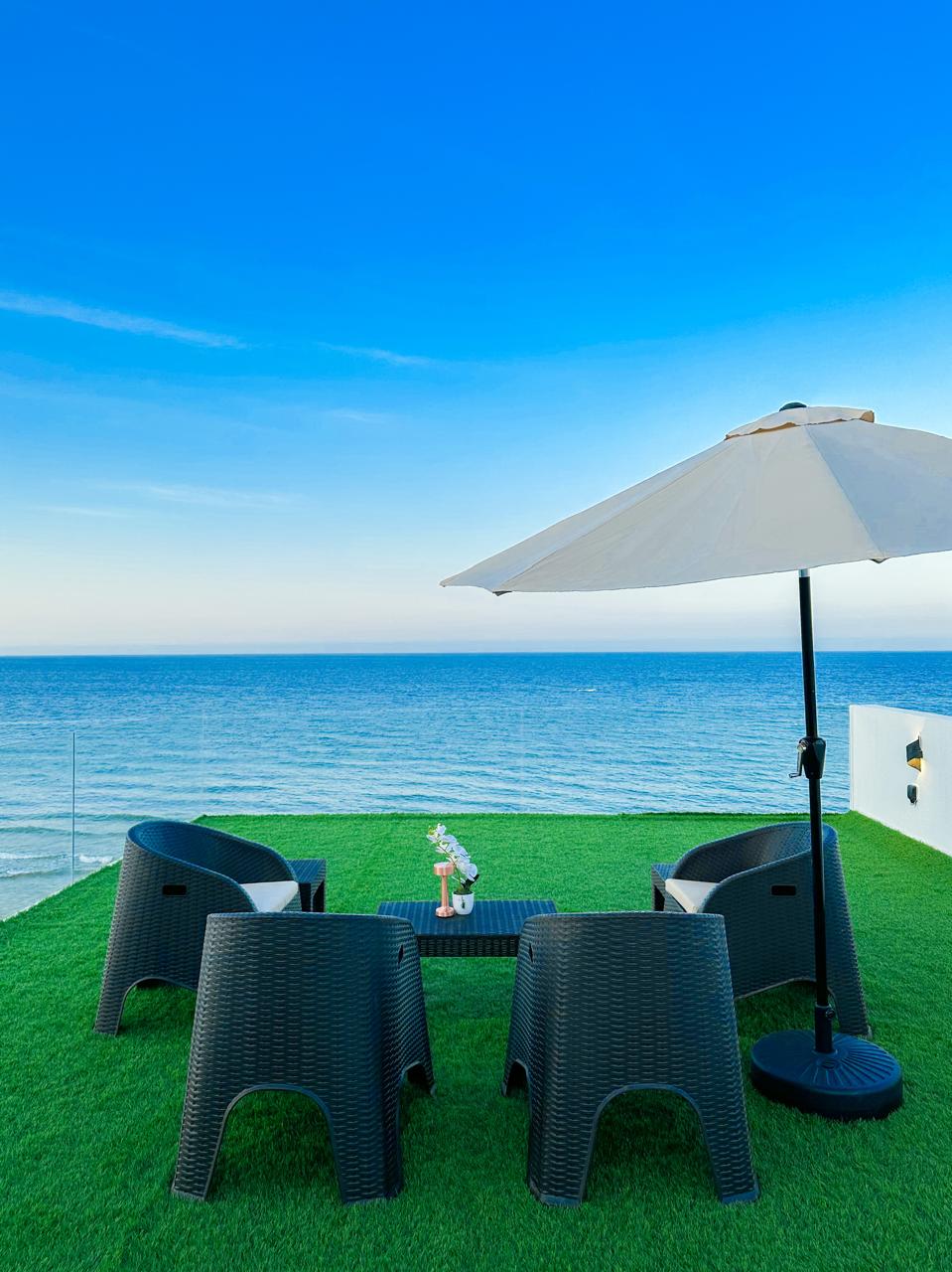 An outdoor seating area features four wicker chairs arranged around a small table, complemented by a large umbrella. The setting overlooks the sea, with a clear blue sky above. The lush green grass enhances the relaxing atmosphere.