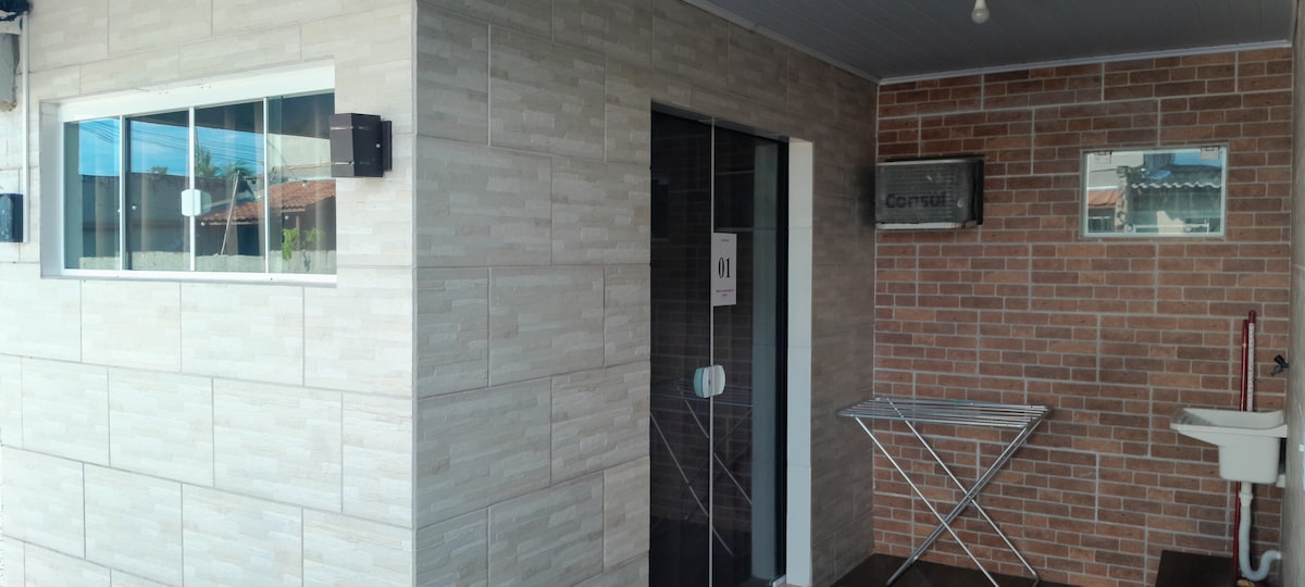 A modern entrance area is presented, featuring a tiled wall adjacent to a brick façade. To the right, a drying rack is positioned beside a small sink. Natural light streams through the large window, offering views of the surrounding outdoor space.