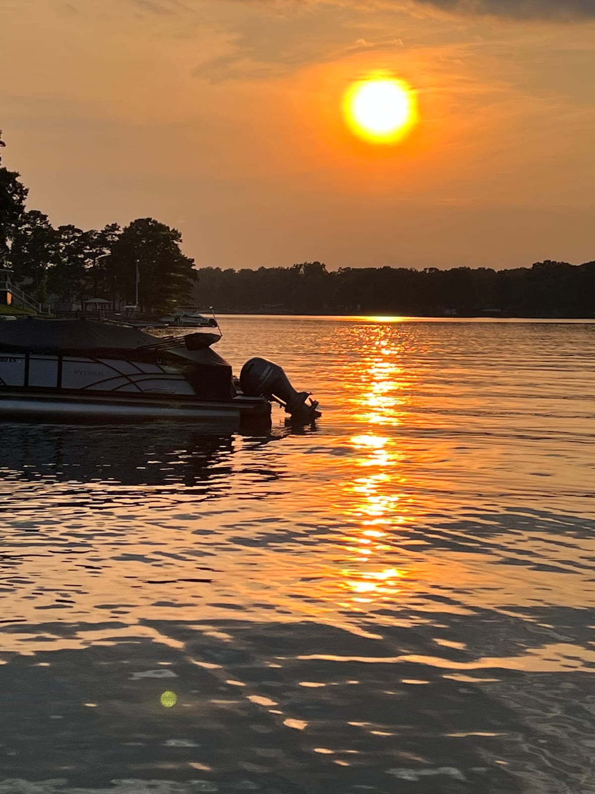 The sun sets over a calm lake, casting golden reflections on the water's surface. A boat is moored nearby, silhouetted against the vibrant sky. Trees line the shore, contributing to the serene lakeside environment.