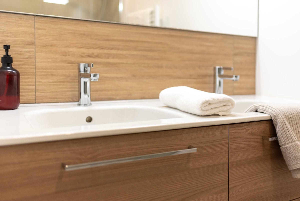 A modern bathroom is featured with a sleek countertop and a well-lit mirror. Two chrome faucets are positioned above a clean sink. A rolled white towel rests beside a beige towel, both neatly placed near the edge of the countertop.