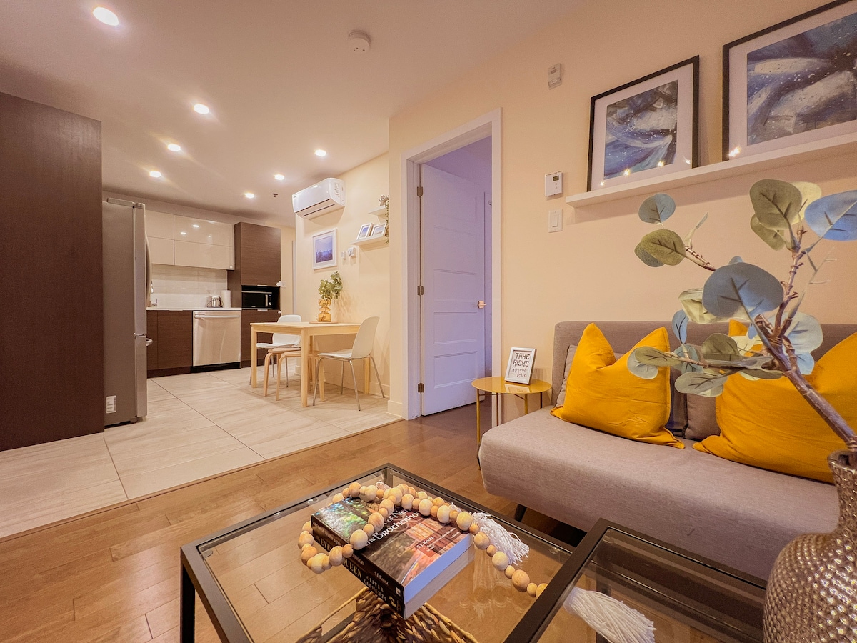 A welcoming living space is presented with a cozy gray sofa adorned with bright yellow pillows. A coffee table holds decorative items, and a dining area with light wood furniture is visible in the background, beside a fully equipped kitchen. Soft lighting enhances the warmth of the room.