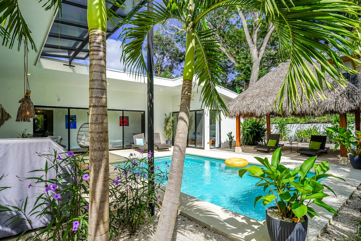 A refreshing pool is framed by lush greenery and tropical plants, featuring a thatched-roof gazebo nearby. Sun loungers are arranged around the pool area, inviting relaxation under the sun. Large glass doors open from the villa, enhancing the connection between indoor and outdoor spaces.