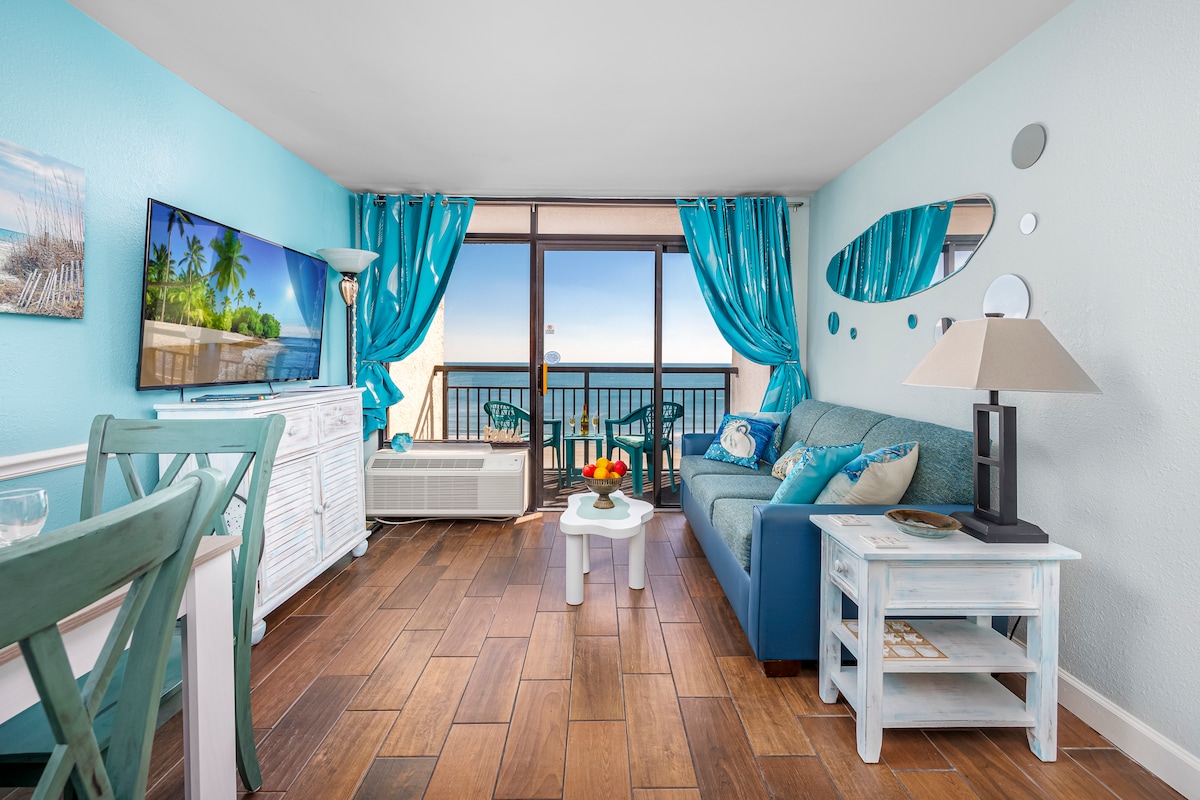 The living area features a comfortable couch adorned with coastal decor, facing a large window that offers ocean views. A smart TV is mounted on the wall, and the space is complemented by turquoise curtains and light wooden accents. A dining area and side table add functionality.