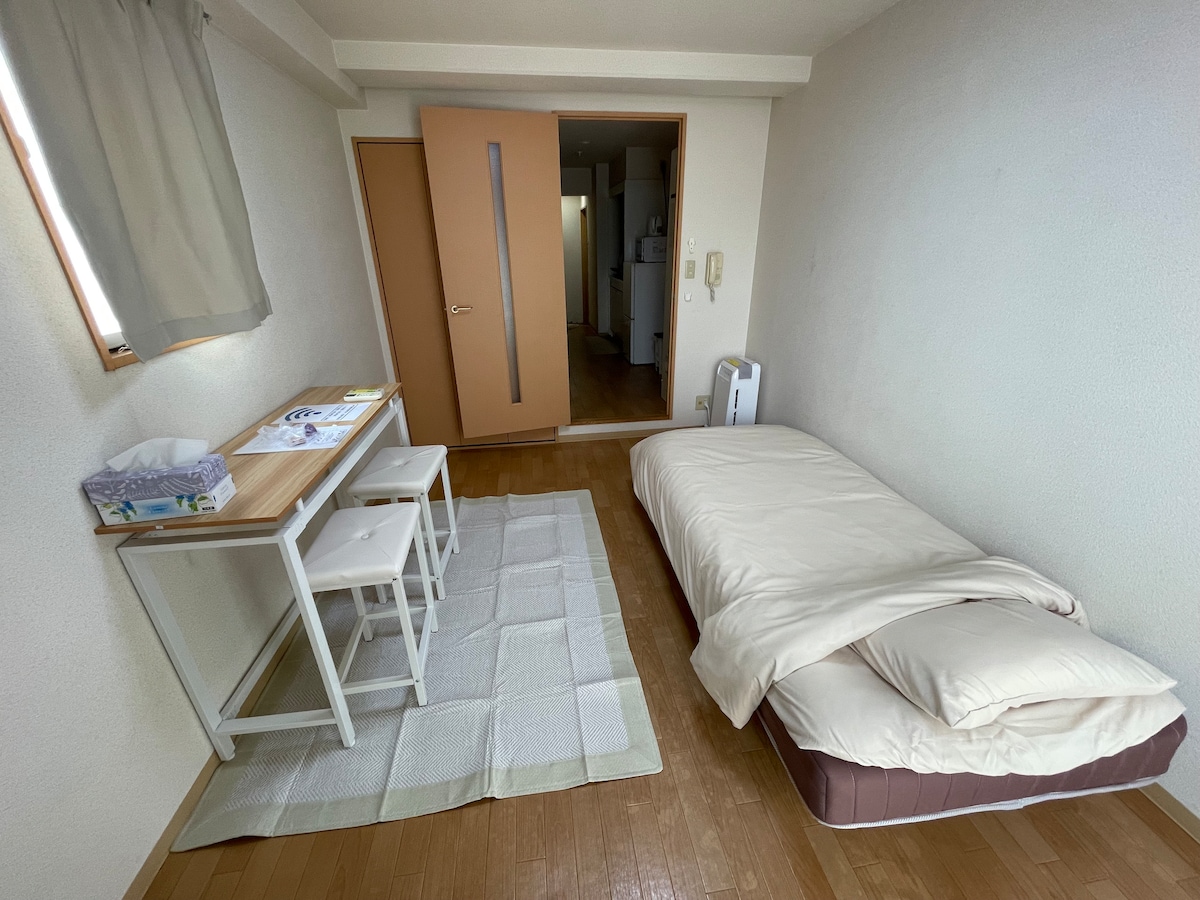 A simple room is equipped with a spacious bed and a small dining area featuring a wooden table and two chairs. A light-colored rug is placed on the wooden floor, and the natural light enters through a window dressed with beige curtains.