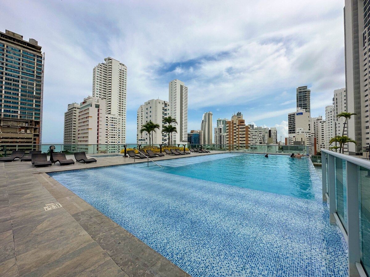 An expansive pool is set atop a high-rise building, surrounded by lounge chairs. The water reflects the sky, while city skyscrapers can be seen in the background. Tropical trees provide a touch of greenery, contributing to a refreshing atmosphere.