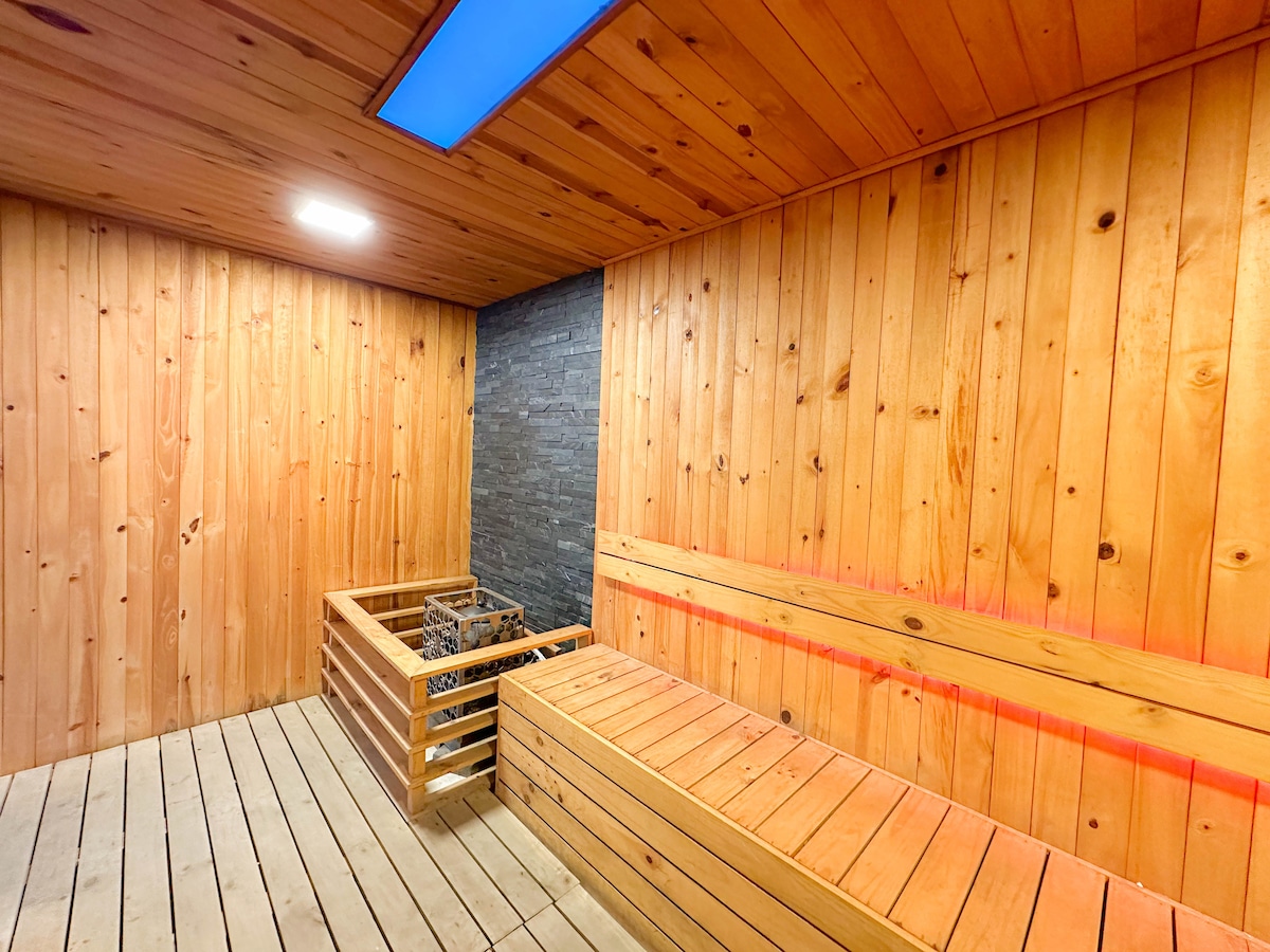 A wooden sauna space features natural wood paneling, creating a warm atmosphere. A bench lines one side, and a stone wall adds a textured contrast. Soft lighting from the ceiling enhances the serene environment, inviting relaxation after a long day.