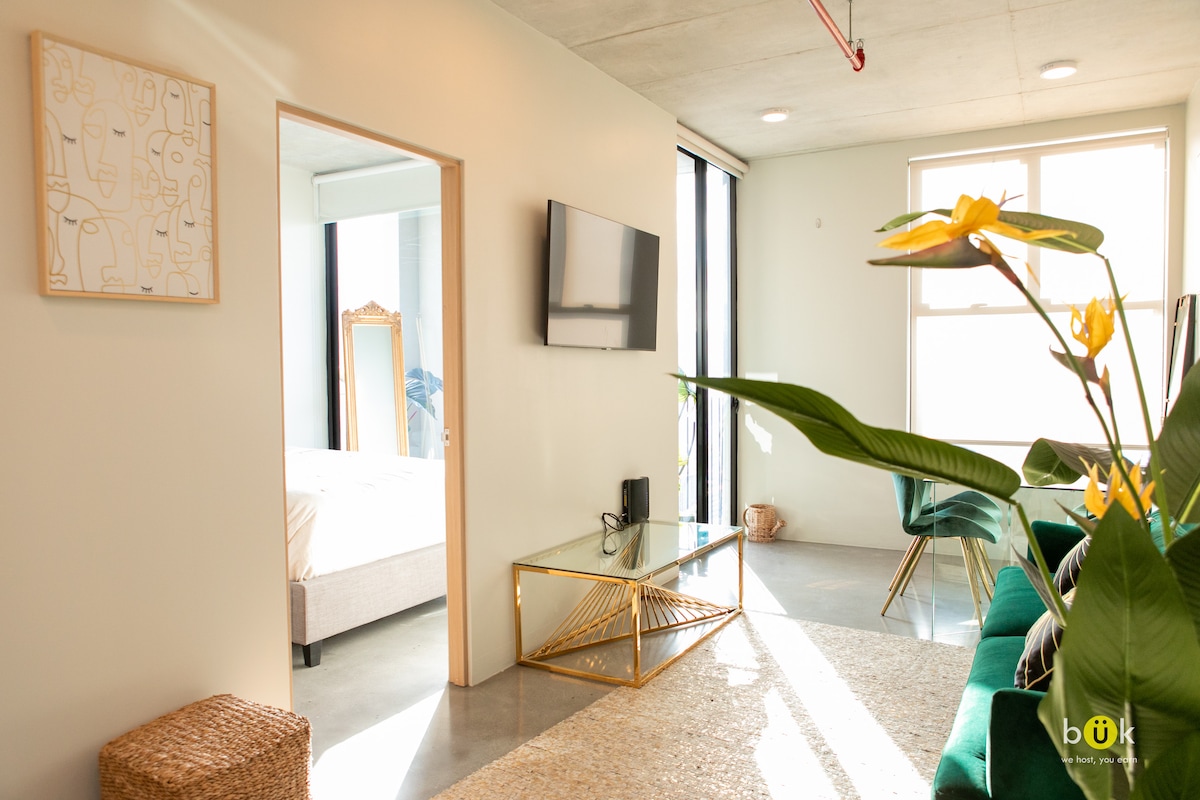 A modern living space is presented with sleek decor, featuring a large mirror and a flat-screen television mounted on the wall. Natural light filters through expansive windows, illuminating the green seating area and the abstract wall art. A decorative plant adds a touch of freshness.