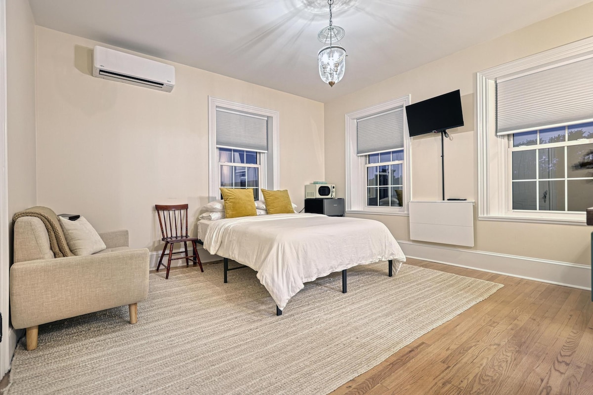 A spacious suite features a queen bed dressed with neutral linens and two decorative pillows. A small loveseat and chair provide seating options. A flat-screen television and a microwave are mounted on the wall. Large windows allow natural light to fill the room.