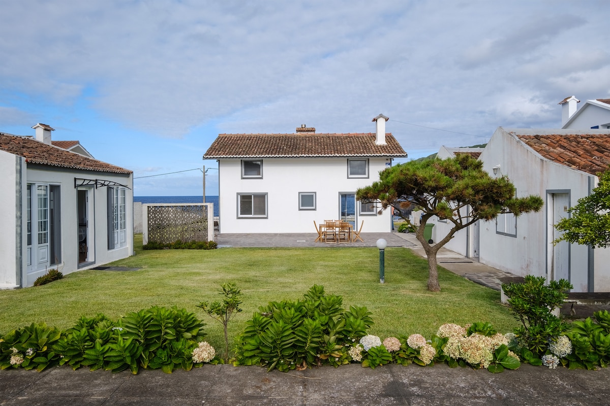 A well-maintained garden surrounds a two-story white house with a brown tiled roof. A wooden dining table and chairs are visible on the patio, offering ocean views. Flower beds with various plants, including hydrangeas, line the edges of the lawn, under a partly cloudy sky.