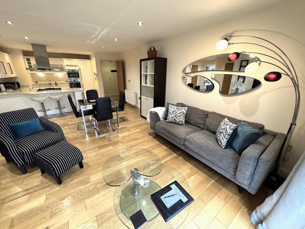 The open-plan living area features a sleek grey sofa accented with plush cushions, a modern glass coffee table, and stylish dining chairs. A striking arc floor lamp and a large wall mirror enhance the space, while warm wood flooring adds a touch of elegance.