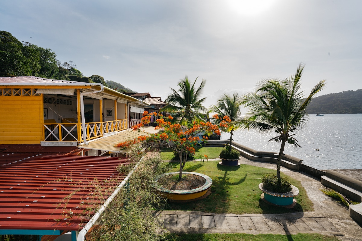 A view of the colorful exterior of the building, featuring yellow walls and a red roof. Palm trees are positioned around a landscaped area with vibrant orange flowers, offering a serene proximity to the bay and a sunlit atmosphere.