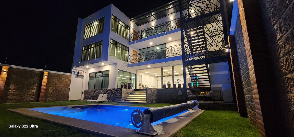A modern multi-story home is showcased at night, with illuminated windows and a well-lit pool area. The spacious outdoor space features a pool, manicured grass, and decorative plants lining the entrance, creating a welcoming ambiance for evening relaxation.