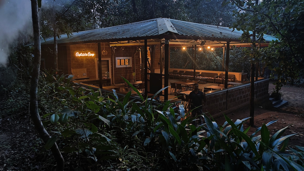The covered outdoor dining area is surrounded by lush greenery, showcasing a rustic design with a metal roof. Soft lighting illuminates the space, highlighting wooden furniture arranged for group gatherings, creating a welcoming environment for dining amidst nature.