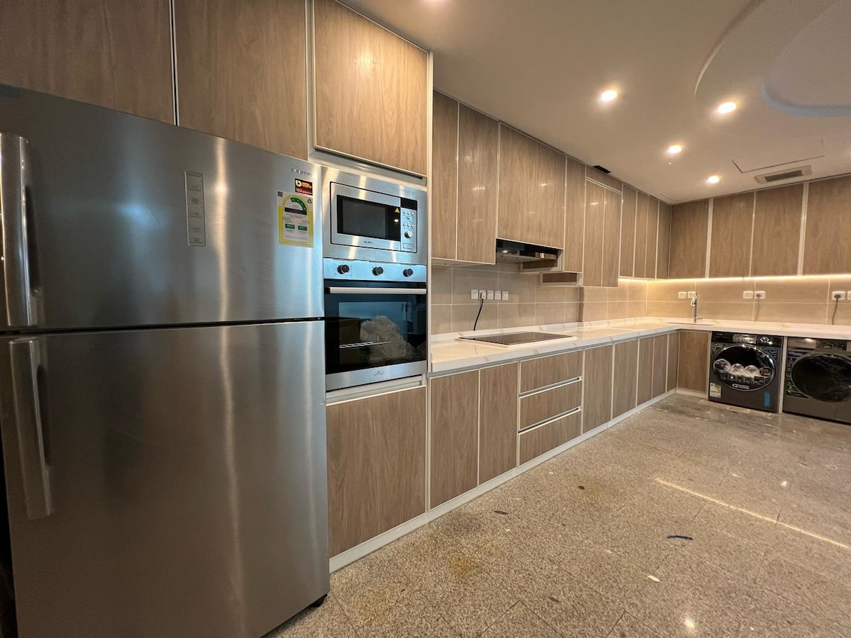 A modern kitchen features sleek wooden cabinetry and stainless steel appliances. A large refrigerator, an oven, and a microwave are included, along with a spacious countertop. Adjacent, a washing machine and dryer are visible, highlighting convenience for guests.