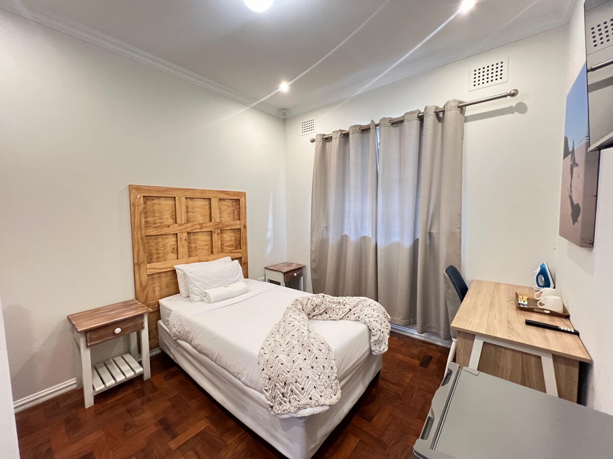 A cozy room features a double bed with a wooden headboard and a light blanket. The space includes a small bedside table and a desk with a chair, while natural light enters through the window, which is covered by light gray curtains.